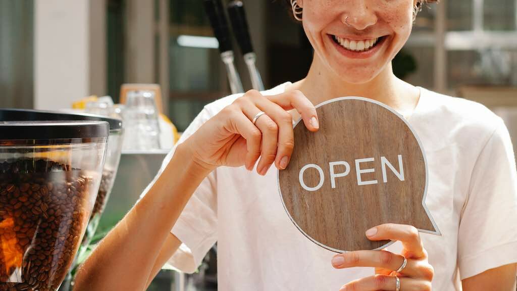 Image of a woman with an 'open' sign. Accompanies the Style Factory resource on how to change your Shopify store name.
